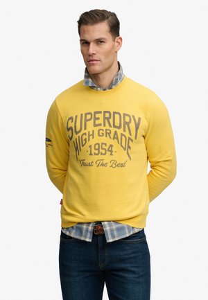 Man draagt een gele Superdry trui over een blauw geruit overhemd en donkere jeans, staand met de handen achter de rug op een witte achtergrond.