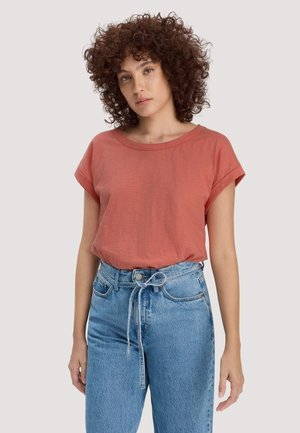 Frau mit lockigem Haar, die ein rostfarbenes T-Shirt mit hochgekrempelten Ärmeln trägt, das in eine hoch taillierte blaue Jeans gesteckt und vorne mit einer Schnur gebunden ist.