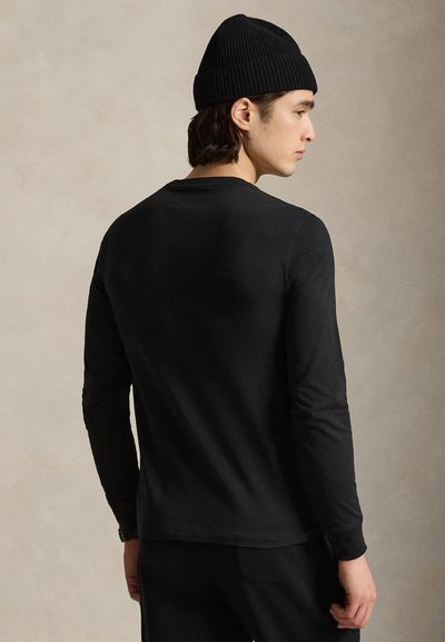 Mężczyzna w czarnej koszuli z długim rękawem, czarnych spodniach i czarnej dzianej czapce, z profilu na neutralnym tle.