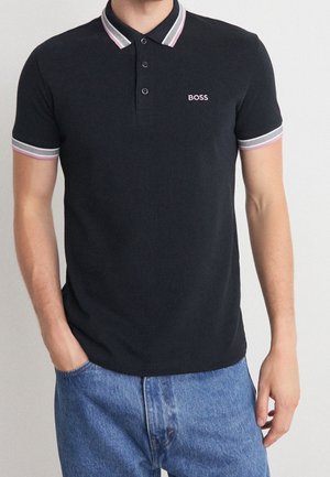 Mies, jolla on musta lyhythihainen poolopaita, jossa on vaaleanpunainen ja valkoinen reunus sekä "BOSS"-logo, yhdistettynä sinisiin farkkuihin, kädet rennosti sivuilla.