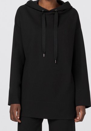 Persoon die een effen zwarte capuchontrui met lange mouwen en zwarte trekkoorden draagt, staand tegen een lichte achtergrond.