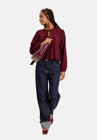 Haut péplum bourgogne à manches longues avec détail à nouer, associé à un jean large bleu foncé et des chaussures rouges ; le mannequin porte un sac à bandoulière bourgogne.