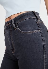 Gros plan d'une personne portant un jean taille haute délavé foncé avec des surpiqûres orange et un top court blanc côtelé.