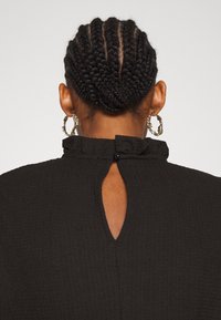 Indumento nero con texture, con scollatura arricciata e un'apertura a forma di chiave sul retro, dotato di chiusura a bottone. Orecchini ad anello argentati in vista.