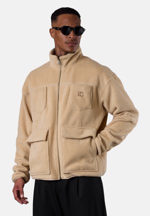 Beige fleece jacket featuring a high collar, front zip closure, and multiple pockets. Soft texture with discreet logo detail on chest.