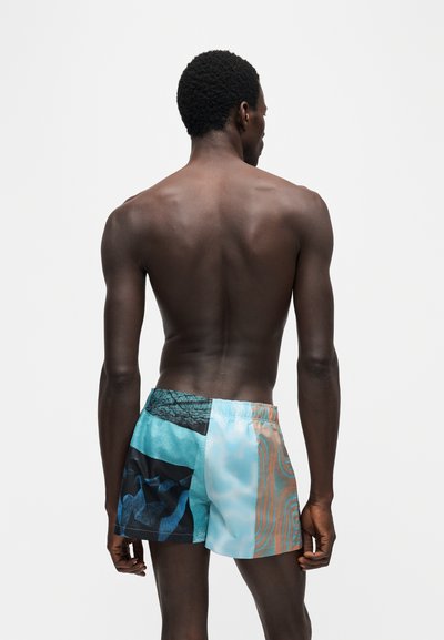 Homme torse nu debout, dos à la caméra, portant un short de bain coloré à motifs bleu, noir et orange, sur un fond uni.
