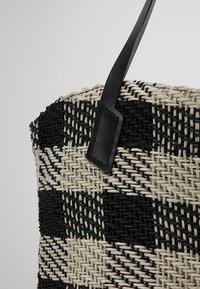 Schwarz- und beige geflochtene Tragetasche mit einem diagonalen karierten Muster und einem schwarzen Ledergriff, der mit einer genähten Lasche befestigt ist.