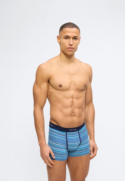 Homme musclé torse nu debout, portant un boxer rayé bleu avec une ceinture de taille bleu marine et un logo de marque sur un fond clair uni.
