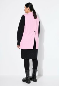 Rosa ärmellose Weste mit Rückenschlitz und Knopfdetails, über einem schwarzen glitzernden Langarmkleid getragen und kombiniert mit schwarzen Knöchelboots.