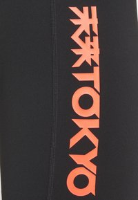 Black athletic leggings featuring bold, orange "TOKYO" text and Japanese characters along the side seam, with a smooth, stretchy texture.