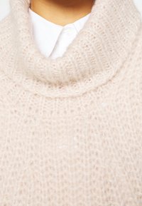 Gestrickter Schalkragenpullover in zartem Beige, mit einer lockeren Webstruktur und Rippmuster. Über einem weißen Hemd mit Kragen getragen.