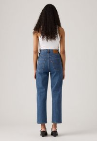 Donna con lunghi capelli ricci, che indossa un top senza maniche bianco, jeans blu a vita alta e scarpe con tacco nere, in piedi di spalle.