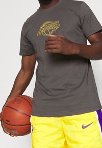 Hombre con camiseta gris de Los Angeles Lakers y pantalones cortos amarillos de los Lakers sosteniendo una pelota de baloncesto contra un fondo blanco.