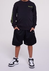 Black sweatshirt with green logo, black mesh shorts, and black and white high-top sneakers. Casual athletic wear with a relaxed fit.