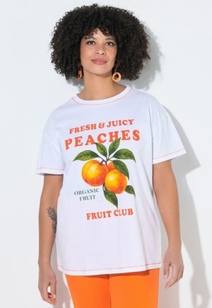 Kobieta o kręconych włosach, ubrana w białą koszulkę z napisem „Fresh & Juicy Peaches” oraz ilustracją brzoskwini, do tego pomarańczowe spodnie i kolczyki.