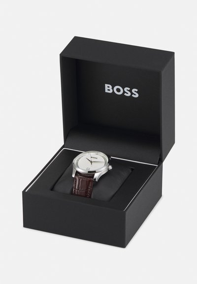 Montre argentée avec un cadran blanc texturé, bracelet en cuir marron et fonction de date, présentée dans une boîte noire portant le logo "BOSS".