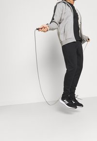 Sudadera gris con cremallera y rayas negras, camiseta negra, pantalones de chándal negros y zapatos negros con suelas blancas, sosteniendo una cuerda para saltar mientras salta.