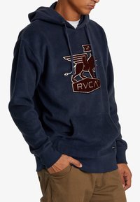 Sudadera de forro polar azul marino con bolsillo frontal, capucha con cordón y logo bordado de un león alado en marrón y blanco. Puños y dobladillo acanalados.