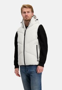 Een lichtgrijze puffervest met een rits aan de voorkant, 2 zijzakken, een capuchon en zwarte accenten, gedragen over een zwart t-shirt met lange mouwen en jeans.