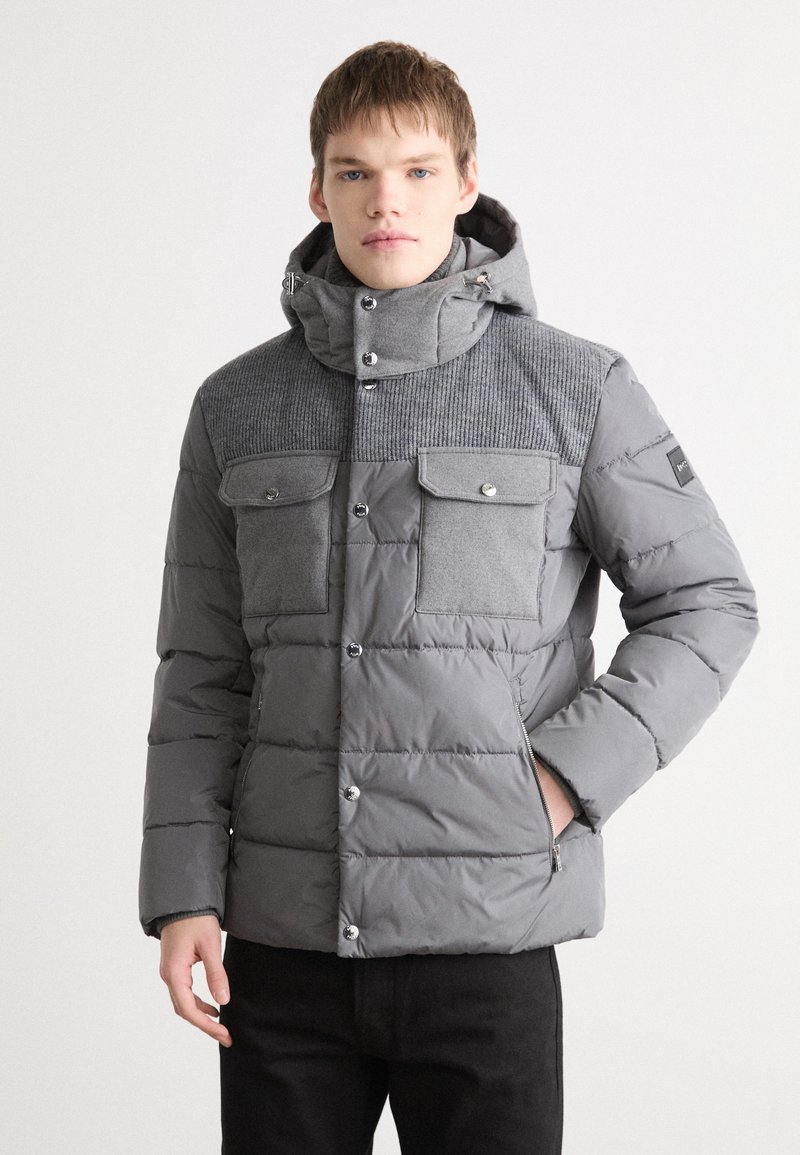 Giacca puffer grigia con parte superiore in tessuto a coste, due tasche sul petto, bottoni a pressione e cappuccio regolabile, con una texture trapuntata su tutta la giacca.