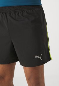 Pantalones cortos deportivos negros con un acabado texturizado, que presentan franjas laterales fluorescentes verdes y un logo de Puma gris en el dobladillo inferior. Cintura elástica.
