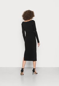 Vestido preto de manga longa com um design ajustado, comprimento a meio da panturrilha e fenda lateral. Combinado com sandálias pretas de salto alto.