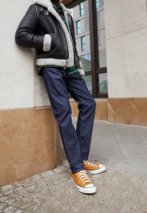 Czarna kurtka wyłożona owczą skórą, zielona i czarna koszula w kratę, niebieskie dżinsy i pomarańczowe niskie trampki z białymi podeszwami, stojąc pod ścianą.