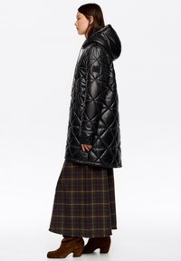 Czarny pikowany puchowy płaszcz z kapturem, z boczną kieszenią na zamek, noszony na brązowej, kraciastej maxi spódnicy i brązowych botkach na obcasie.