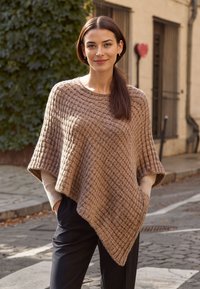 Poncho lavorato a maglia color beige con un motivo a diamante testurizzato; presenta una vestibilità ampia e maniche larghe, indossato sopra pantaloni scuri. Ambientazione all'aperto.