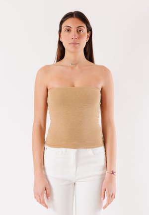 Donna con lunghi capelli scuri che indossa un top beige senza spalline e pantaloni bianchi, in piedi davanti a uno sfondo bianco semplice.