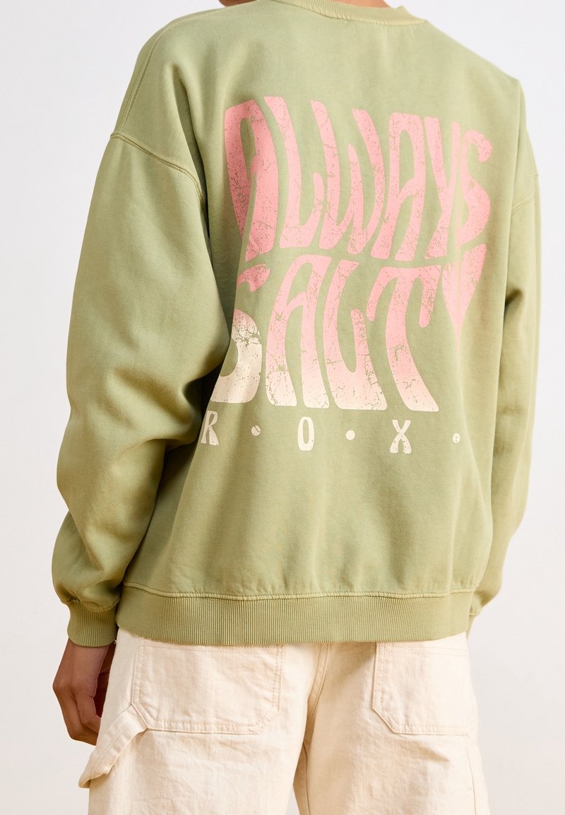 Personne portant un sweat-shirt vert clair avec le texte rose « Always Salty » et le texte blanc « R.O.X. » dans le dos, associé à un pantalon cargo crème.