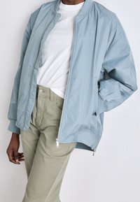 Světle modrá bomber bunda se zipovým zapínáním a žebrovaným límcem. Má boční kapsy a nenápadné logo. Kombinováno s bílým tričkem a zelenými kalhotami.