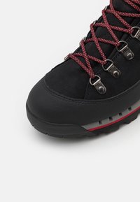 Botas de senderismo negras con una puntera de goma texturizada, cordones rojos y negros, ojales metálicos y una suela con un patrón en negro y gris.
