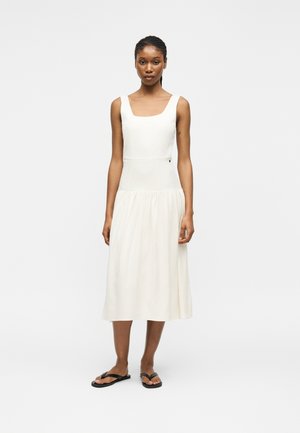 Donna in piedi che indossa un vestito midi bianco senza maniche con scollo quadrato e sandali neri bassi su uno sfondo chiaro uniforme.