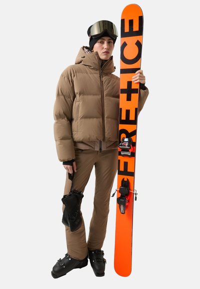 Brązowa kurtka puchowa i spodnie, czarne buty narciarskie, trzymający pomarańczową nartę z czarnym napisem, w okularkach narciarskich i czapce beanie w kolorze czarnym.