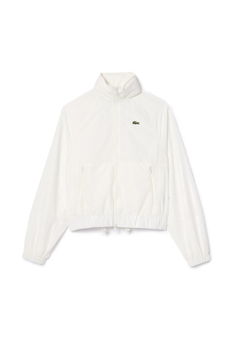 Biała kurtka przeciwwiatrowa z wysokim kołnierzem, zamek błyskawiczny, elastyczne mankiety i dół. Posiada małe zielone logo Lacoste na klatce piersiowej.