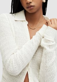 Cardigan bianco e testurizzato con un motivo ondulato, caratterizzato da un collare a punta e chiusura a bottoni. Accentuato da una delicata collana con perle dorate rotonde.