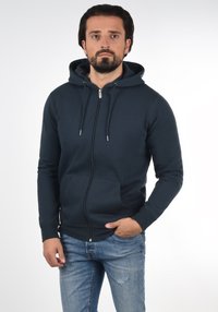 Marineblaues Zip-Hoodie mit Kordelzug, vordere Kängurutasche und gerippte Bündchen. Kombiniert mit hellblauen, distressed Jeans.