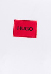 Biała bawełniana koszulka z czerwonym prostokątnym naszywką z napisem "HUGO" w czarnym nadruku. Gładka tekstura, casualowy design.