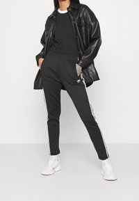 Černé sportovní kalhoty s bílými bočními pruhy, zkombinované s černým tričkem a černou bundou z umělé kůže. Vzhled doplňují bílé sportovní boty.