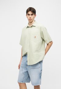 Νεαρός άνδρας με ανοιχτό πράσινο υφασμάτινο κοντομάνικο πουκάμισο και χαλαρά ανοιχτό μπλε τζιν σορτς, μπροστά σε απλό λευκό φόντο.