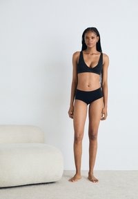 Black bralette and bikini set, seamless design, soft fabric, minimalistic style, fitted cut, shown against a light background and textured floor.