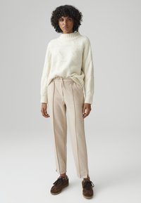 Gebreide crèmekleurige trui met hoge hals en gestructureerd patroon. Lichtbeige broek met plooien, taps toelopende benen en bruine suède schoenen.