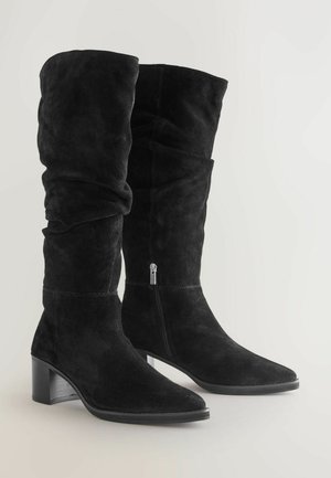 Juoda suedinė, iki kelių siekanti batų pora su laisvu dizainu, smailiu priekiu, blokiniu kulnu ir šoniniu užtrauktuku. Lygi tekstūra su matinio paviršiumi.