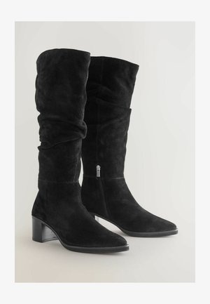 Fekete velúr, térd fölé érő csizma, laza formával, hegyes orral, tömbsarkúval és oldalsó cipzár részlettel. Simább textúrájú, matt felülettel.