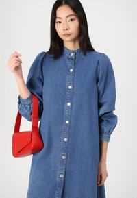 Denim dress with puffed sleeves, button-up front, and a high collar. Paired with a small red handbag. White background.