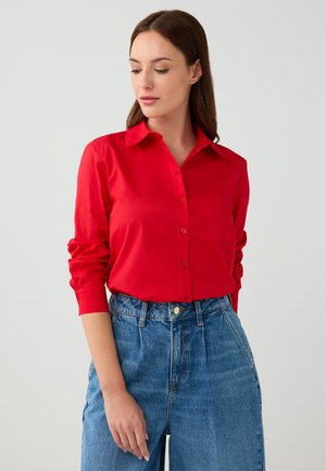 Camicia rossa a maniche lunghe con bottoni, realizzata in tessuto liscio, con colletto a punta e design aderente, abbinata a jeans in denim blu a vita alta.