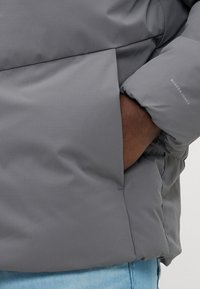 Veste rembourrée grise avec une texture matelassée, dotée d'une poche latérale et de poignets élastiques. Nom de la marque affiché en blanc sur la manche.