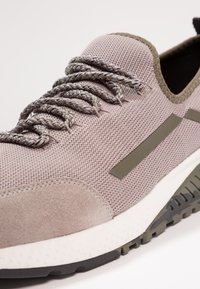 Buty sportowe z jasnofioletową siateczkową cholewką, szaro-zielonymi plecionymi sznurówkami, brązowymi zamszowymi akcentami oraz białą, teksturowaną podeszwą z ciemną gumową bieżnikiem.