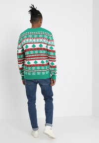 Zielony świąteczny sweter z jaskrawymi, czerwono-białymi wzorami. Dekoracje z płatków śniegu i choinek; ściągacze na mankietach i dole. Noszony z niebieskimi jeansami.